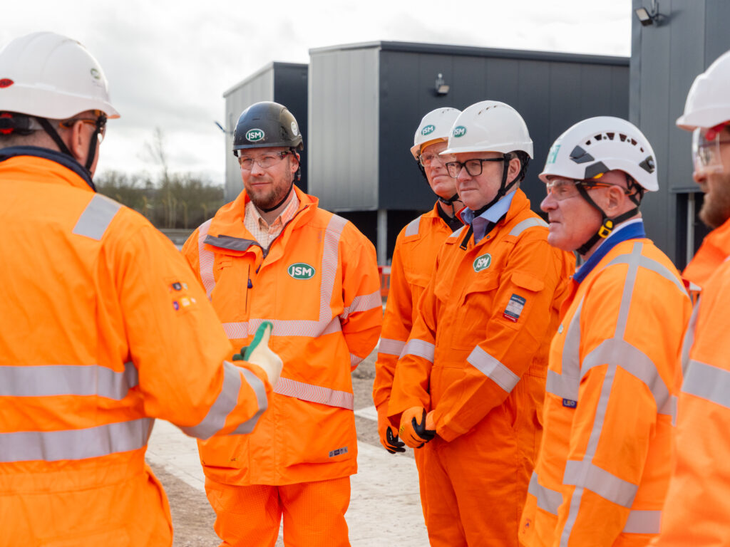 Group of construction workers in bright orange safety gear and white helmets listening to a briefing at a site with equipment in the background, outdoors.