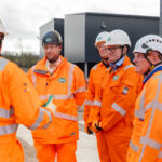 Group of construction workers in bright orange safety gear and white helmets listening to a briefing at a site with equipment in the background, outdoors.