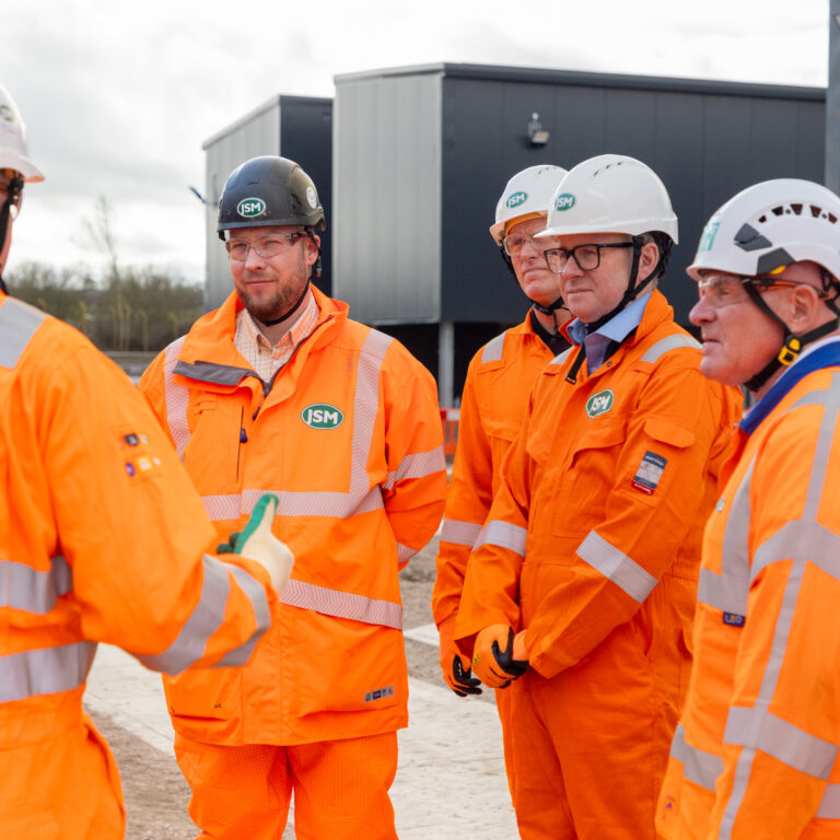 Group of construction workers in bright orange safety gear and white helmets listening to a briefing at a site with equipment in the background, outdoors.