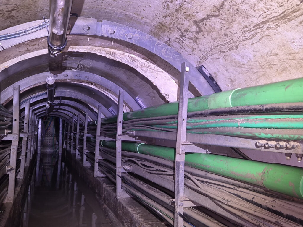 Underground tunnel with bundled green and gray pipes along the curved concrete ceiling and metal supports; a wet floor.