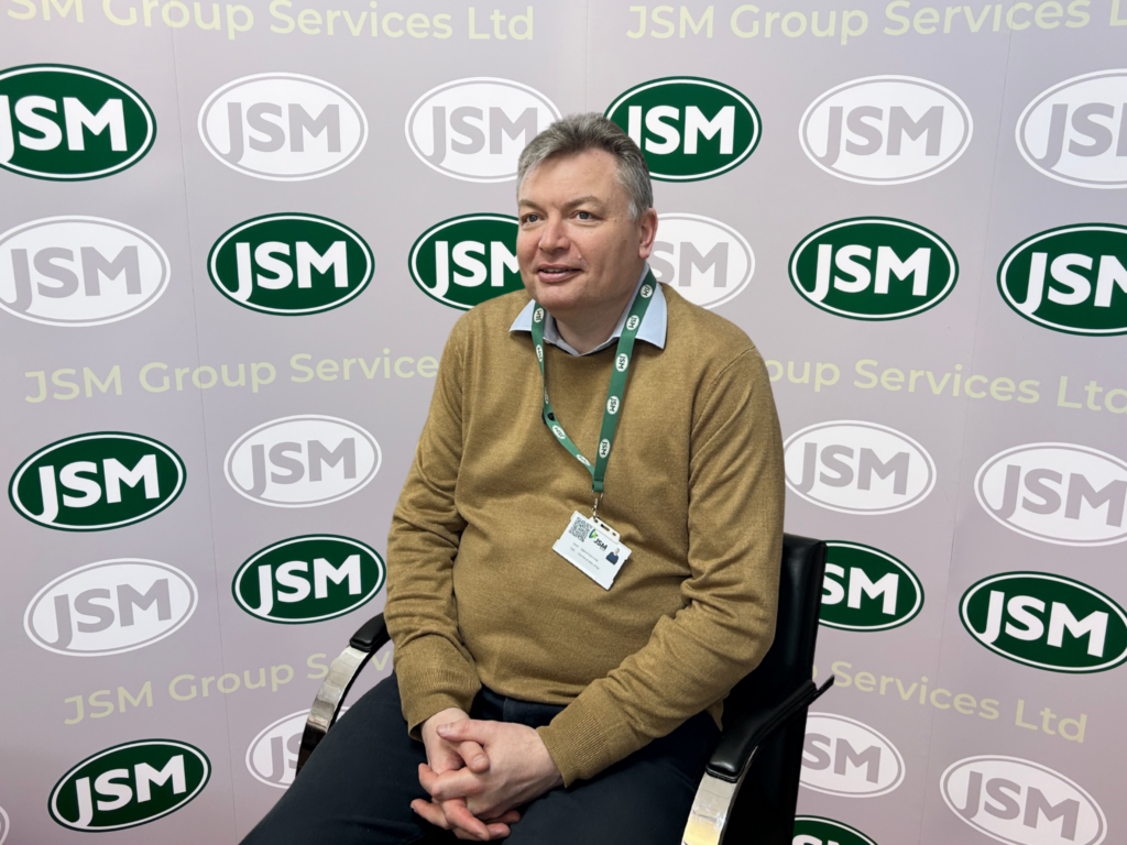 Portrait of a man in a brown sweater wearing a JMS lanyard, seated in front of a JMS backdrop with repeating logos.