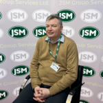 Portrait of a man in a brown sweater wearing a JMS lanyard, seated in front of a JMS backdrop with repeating logos.
