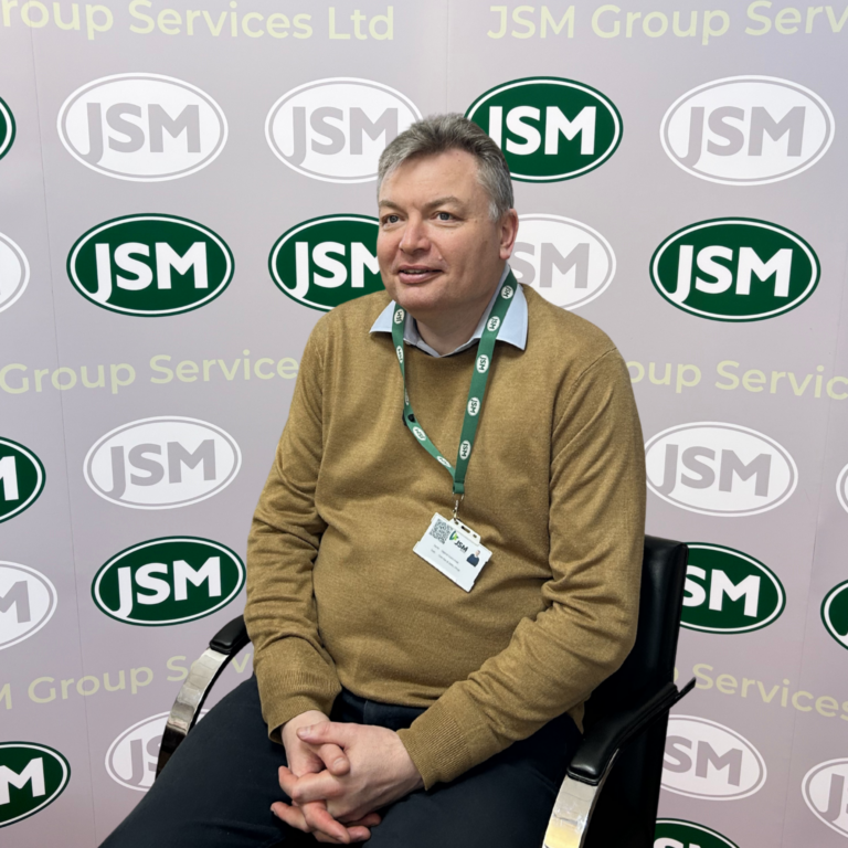 Portrait of a man in a brown sweater wearing a JMS lanyard, seated in front of a JMS backdrop with repeating logos.